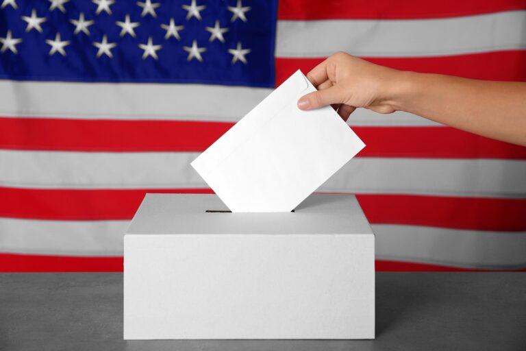 A hand places a white ballot into a plain ballot box in front of an American flag.