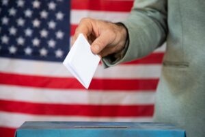 A hand holds a folded white ballot above a plain blue ballot box, with an American flag behind it.