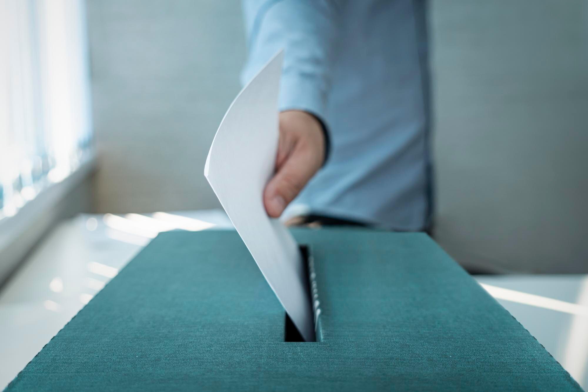 A person places a folded white ballot into a plain teal ballot box.