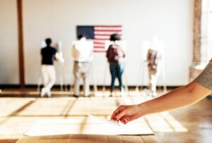A hand places a ballot in a box as people use voting booths beneath an American flag.
