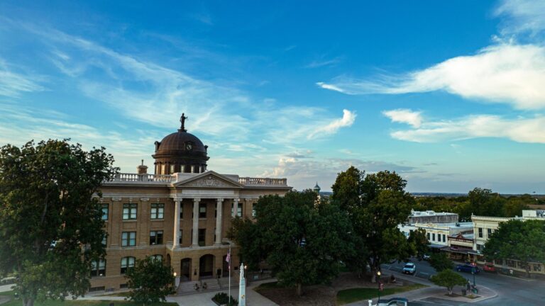 Courthouse framed by trees in Georgetown, Texas, with surrounding streets beneath a blue, cloud-streaked sky.