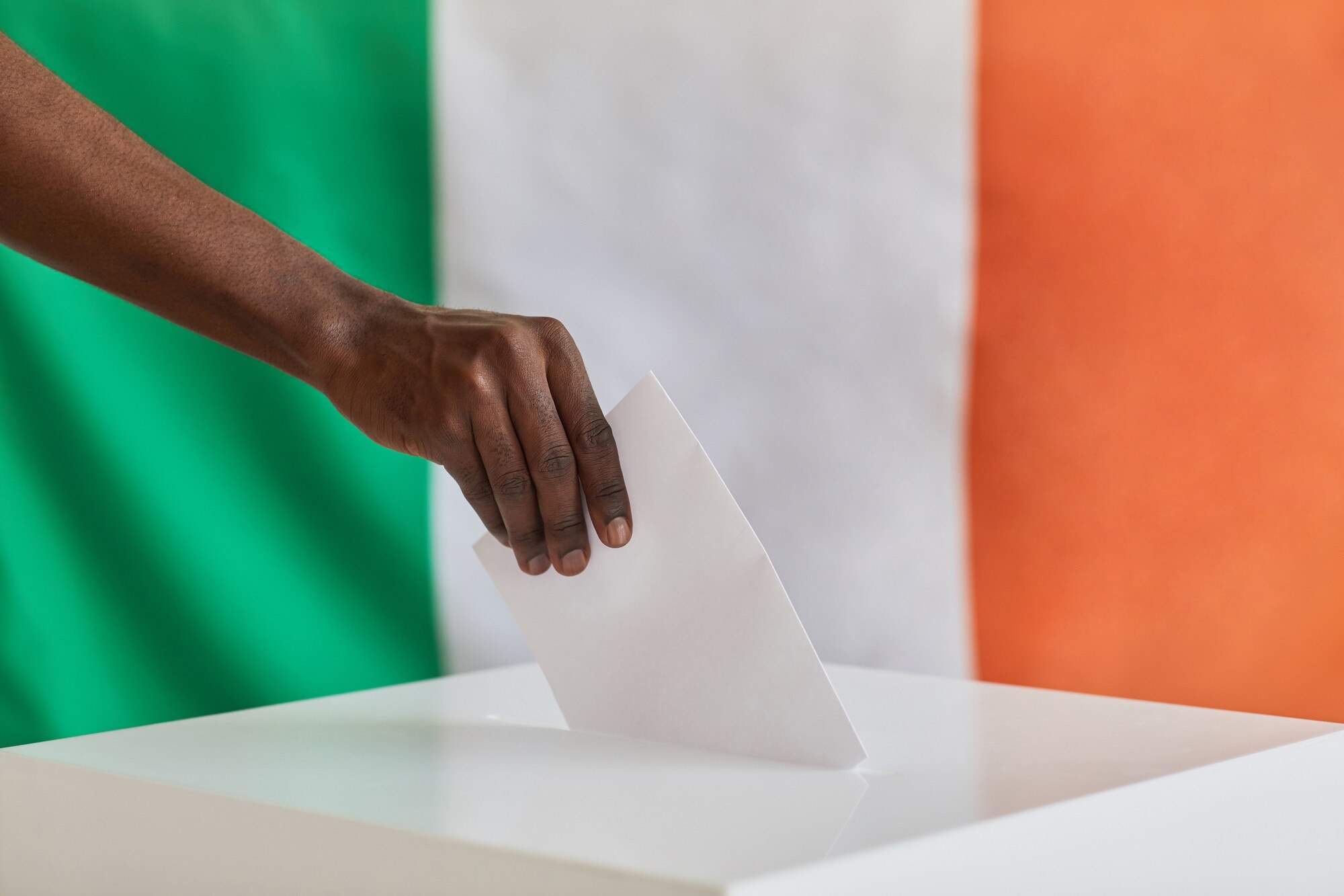 A hand places a white envelope into a ballot box, with a green, white and orange flag behind it.