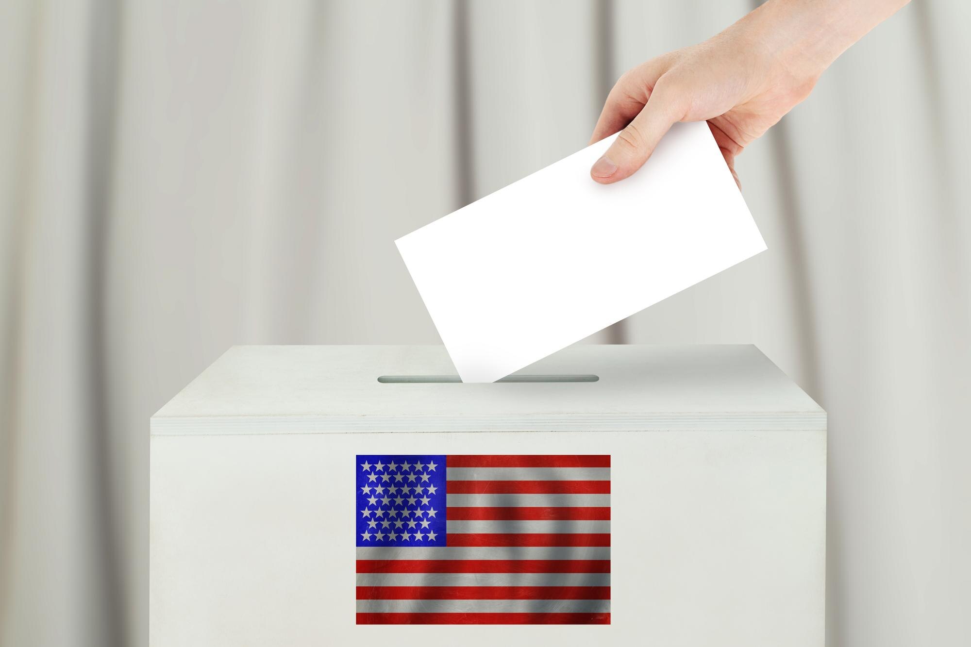 A hand places a blank white ballot into a ballot box decorated with an American flag.