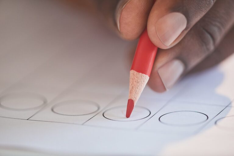 Close-up of a hand holding a red pencil over a circle on a ballot.